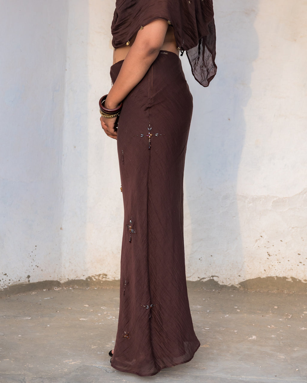 A woman wearing a mocha brown two-piece outfit with a sheer draped dupatta and a long shaped skirt, standing against a plain off-white wall.