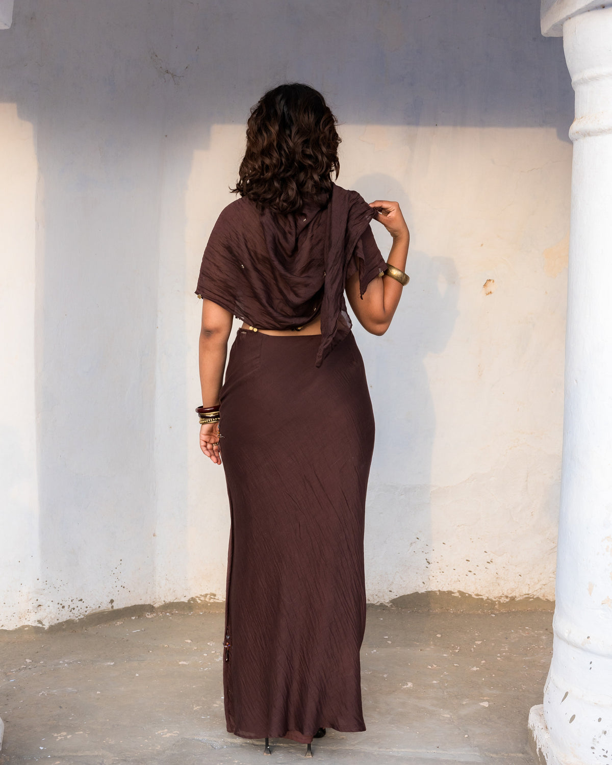 A woman viewed from behind wearing a dark brown semi flared chanderi skirt with intricate metallic handwork and no elastic, standing in a rustic white-walled corridor.