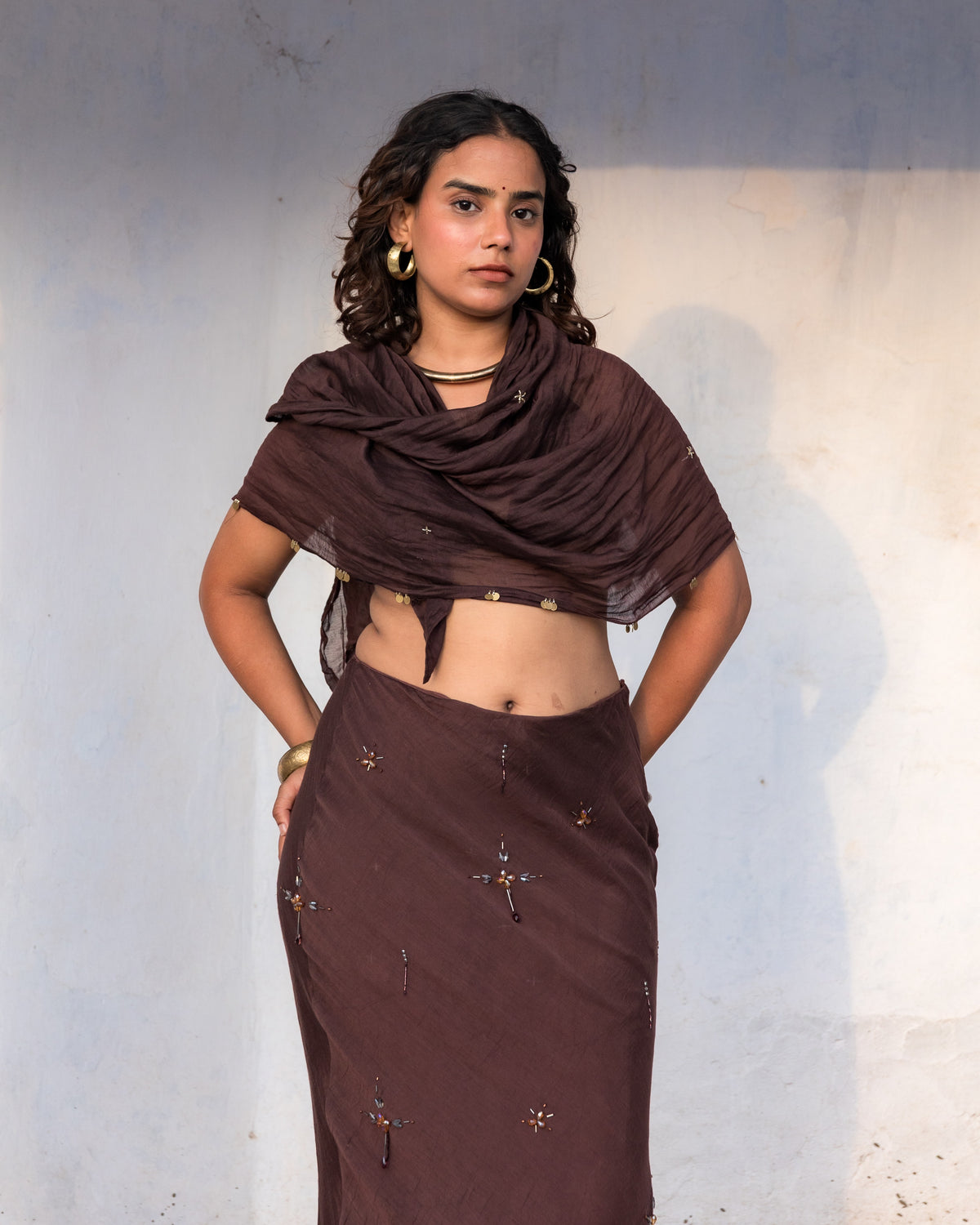 Closeup of a woman modeling a dark brown long skirt with indian beadwork and stonework all handdone, standing against a plain wall.