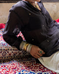 Woman wearing a black long-sleeve embroidered short kurta and white pants, sitting on a red and blue patterned Persian rug. The person is accessorized with multiple silver bangles and a ring.