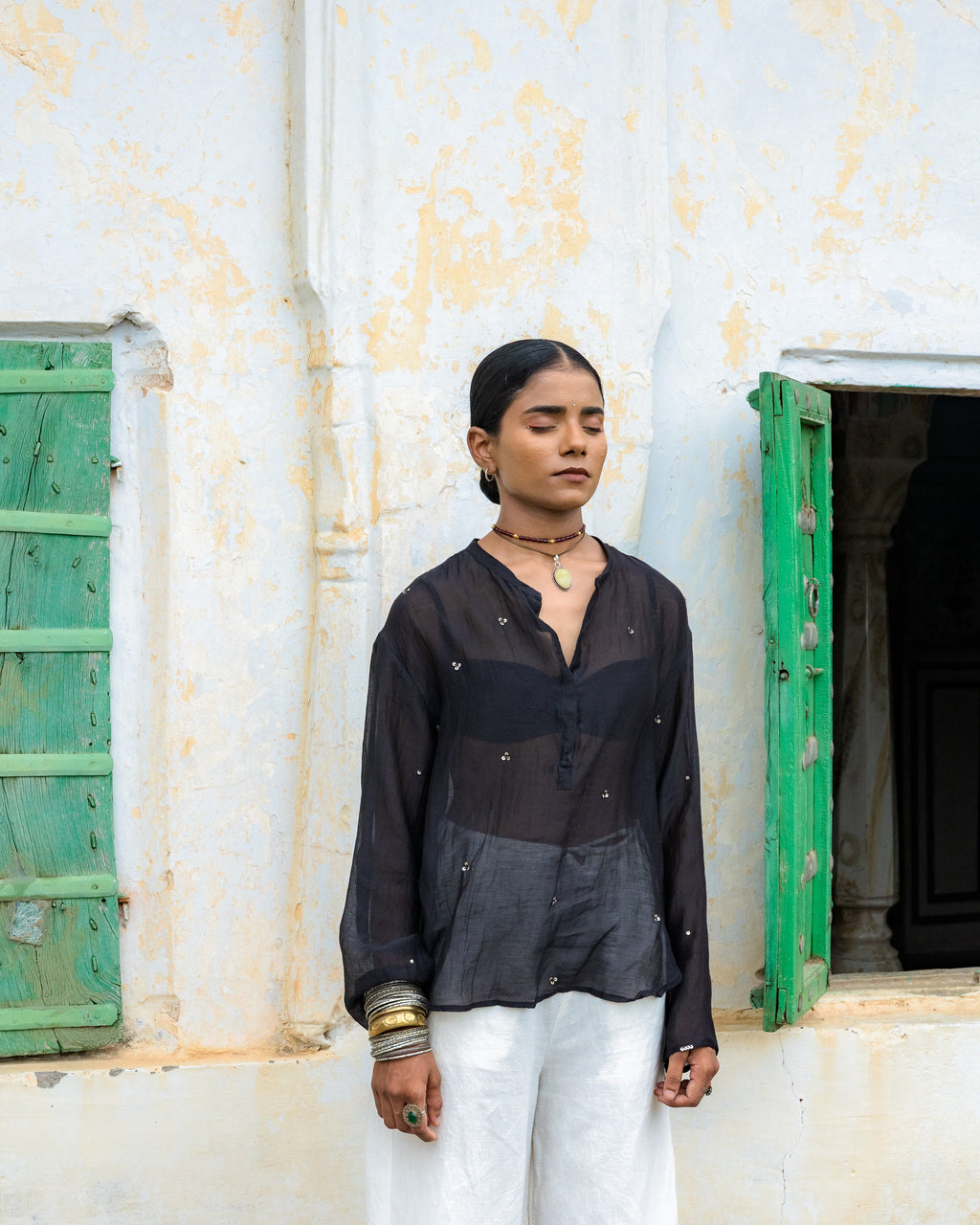 A woman stands against a weathered white wall with green shutters, wearing a black long-sleeve top with metallic embellishments and white wide-leg pants.