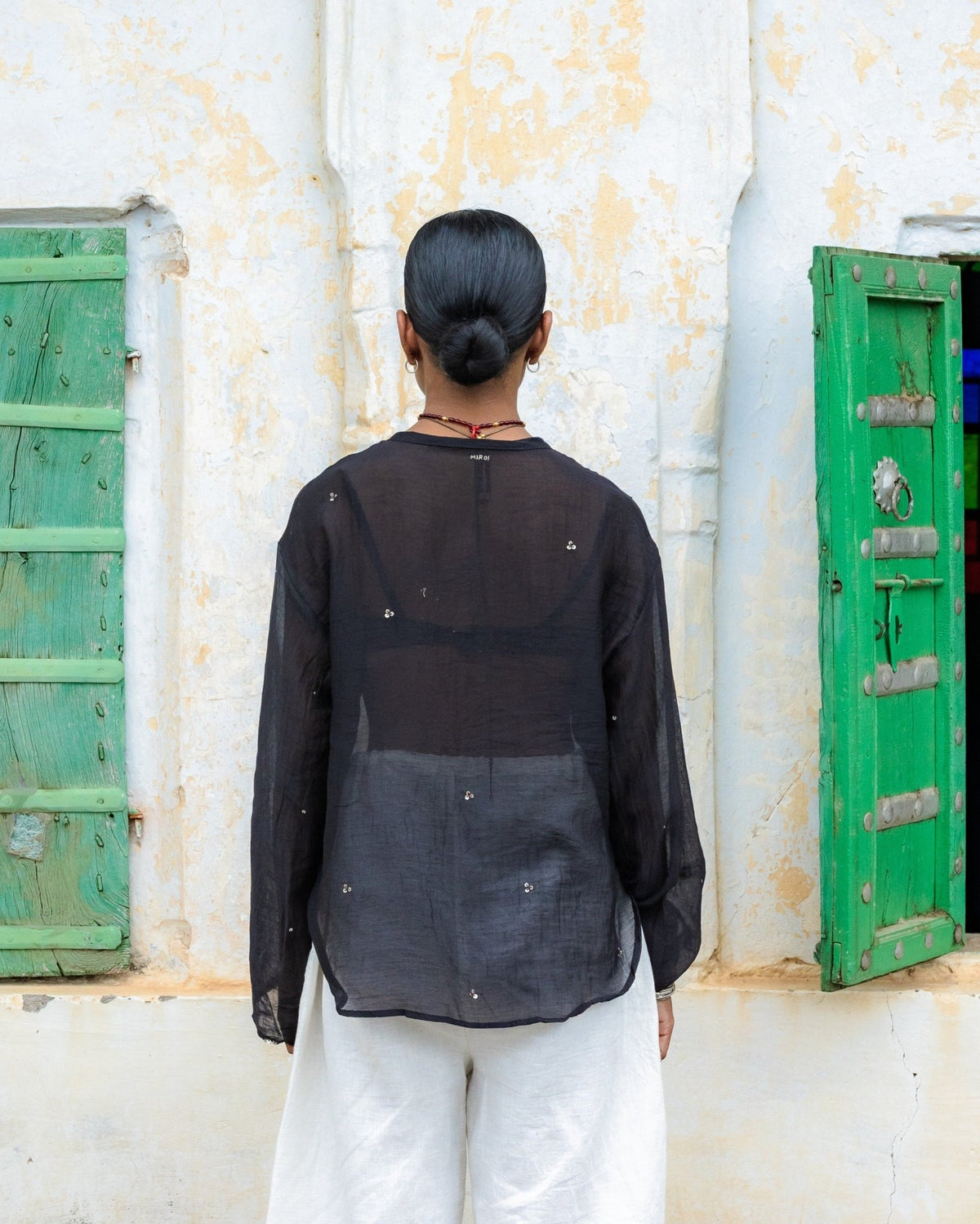 A woman viewed from behind wearing a sheer black long-sleeve brass metal embellished top and white pants, standing in front of a white wall with green shutters.