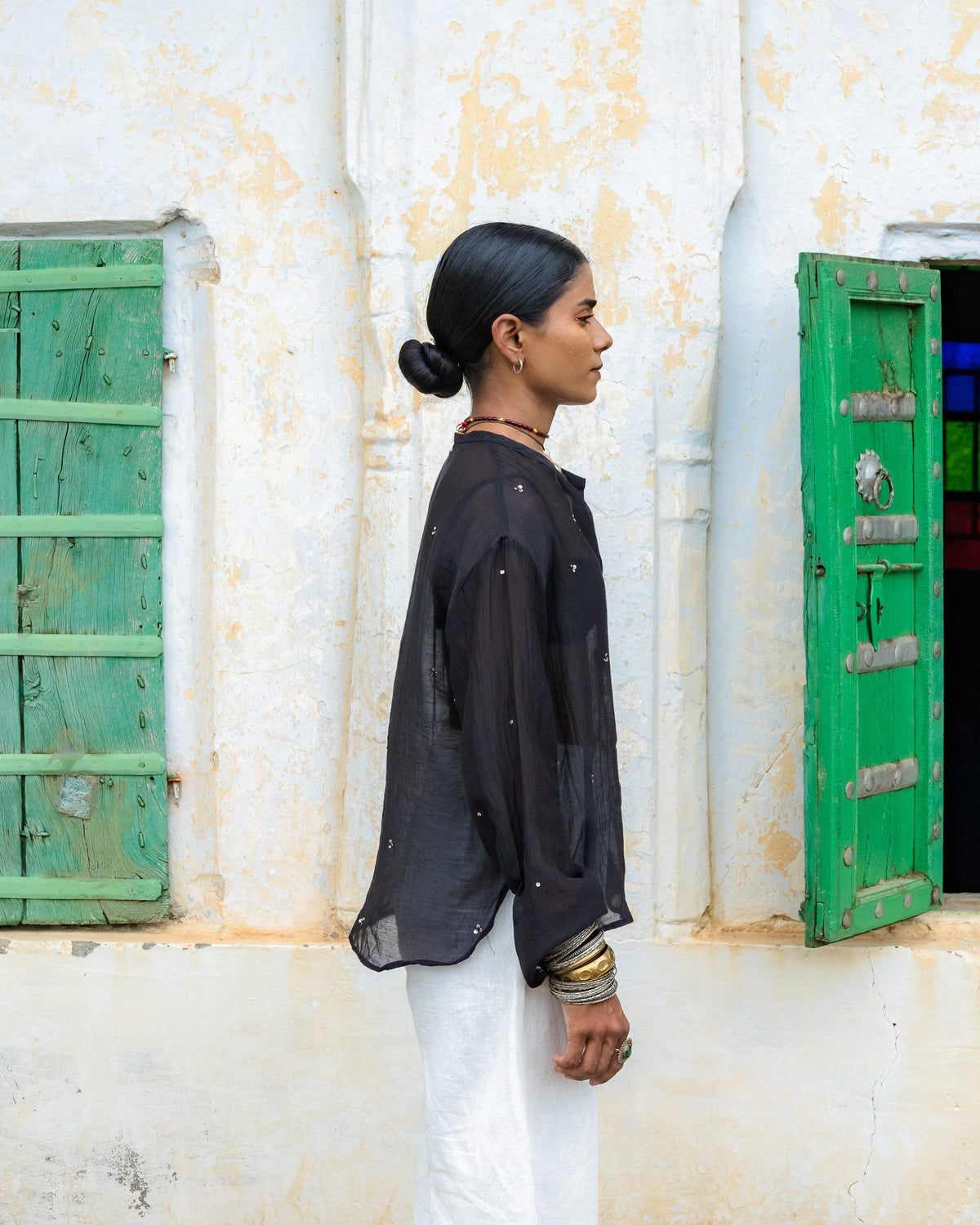 A woman in profile wearing a black long-sleeve semi transparent embroidered top and white pants stands in front of an old white wall with green shutters.