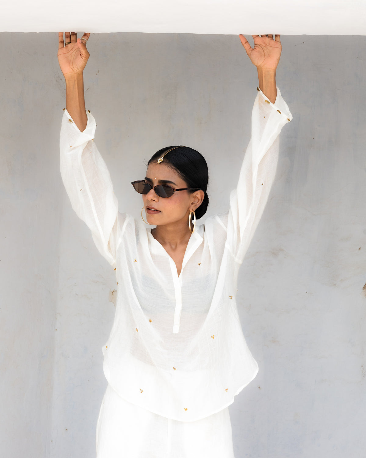 A woman wearing a white long-sleeve short kurta with front placket with small embroidered details and matching white trousers, standing with her arms raised under a white structure against a light gray wall.
