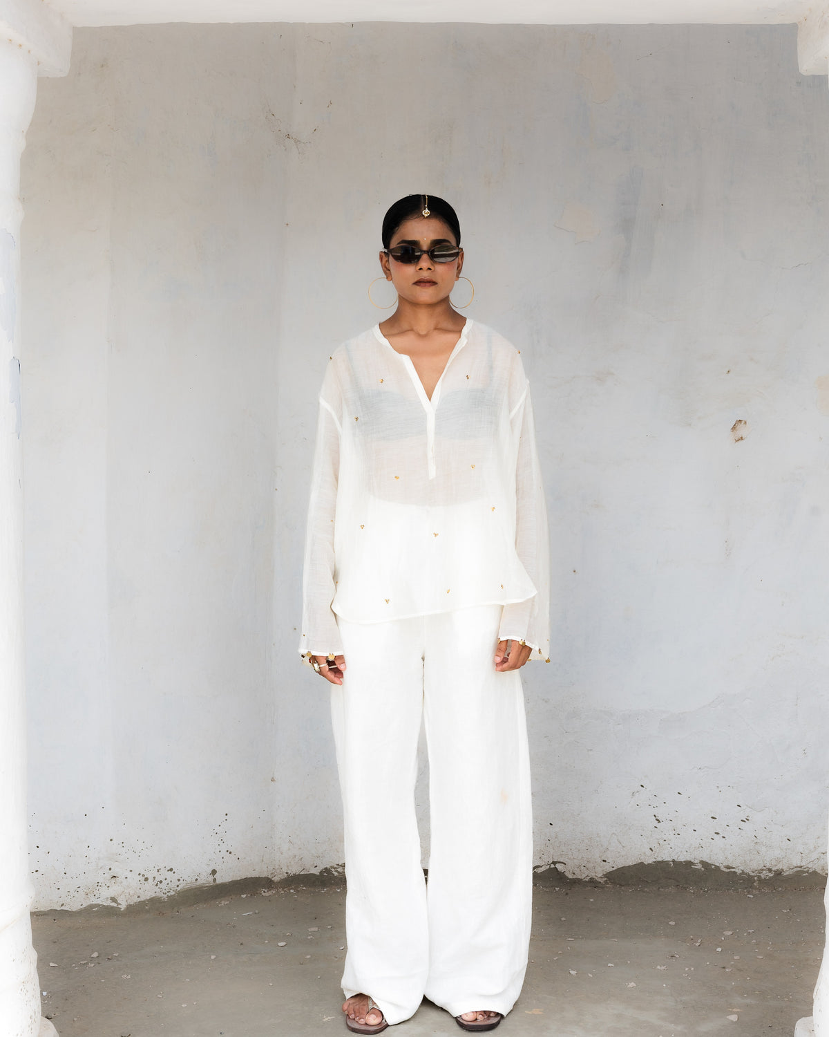 A model stands wearing a white sheer short kurta consisting of a sheer long-sleeve button-front top and matching wide-leg pants.