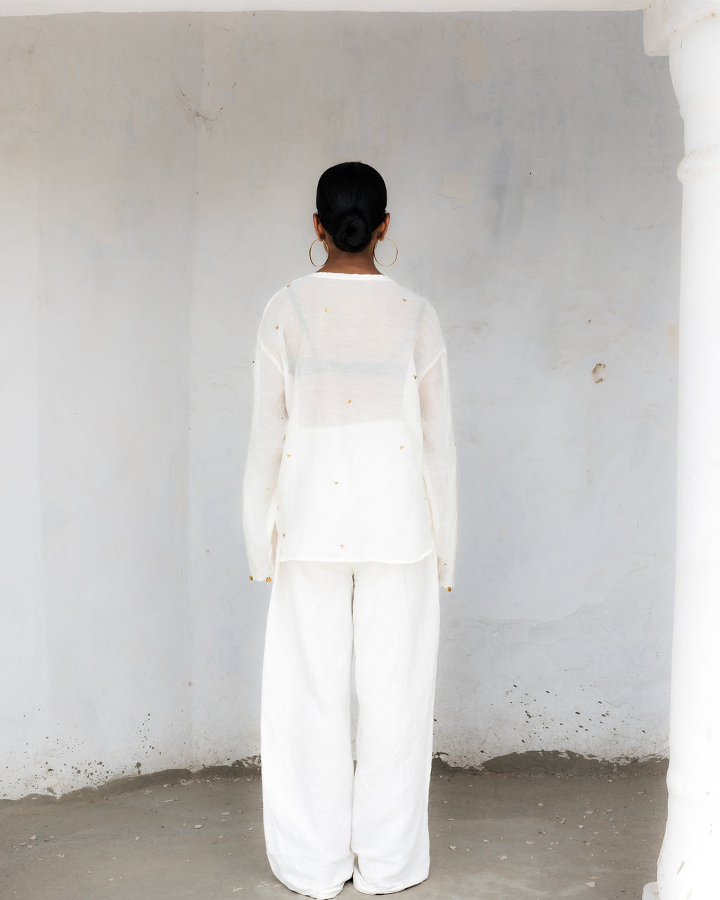 A model viewed from behind wearing a white kurta outfit consisting of a sheer long-sleeve top and wide-leg trousers, standing in a minimalist space.