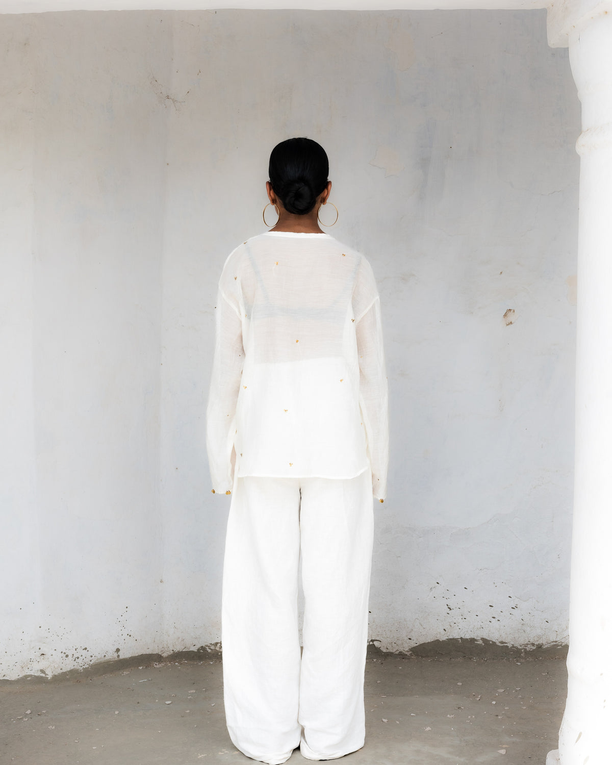 A model viewed from behind wearing a white kurta outfit consisting of a sheer long-sleeve top and wide-leg trousers, standing in a minimalist space.