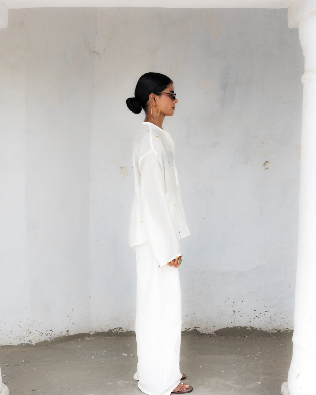 A woman in a side profile wearing a white long-sleeve top, embellished brass sequence, and matching straight pants, standing in a minimalist white-walled architectural setting.